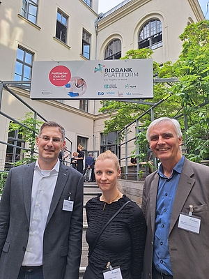 Drei Personen mit Namensschildern stehen vor einem Banner mit der Aufschrift 'Biobank Plattform' und 'Politischer Kick-Off 21 Juni 2023' in einem Innenhof mit Bäumen und Gebäuden im Hintergrund.  Es wurde Text erkannt (OCR). Im Ergebnis wurden Eigennamen ge