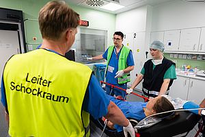 Drei medizinische Fachkräfte in einem Krankenhaus-Schockraum, eine trägt eine gelbe Weste mit der Aufschrift 'Leiter Schockraum', eine Patientin liegt auf einer Liege.
