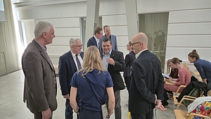Gruppe von Menschen in einem hellen Raum, einige stehen im Kreis und unterhalten sich, andere sitzen auf Stühlen und schreiben oder lesen.