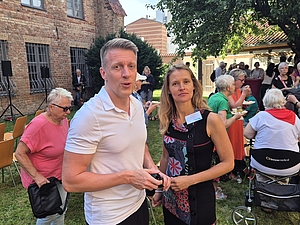Mann in weißem Poloshirt und Frau in schwarzem Kleid mit Blumenmuster stehen im Garten vor einer Gruppe sitzender und stehender Menschen bei einer Veranstaltung