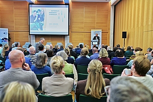 Publikum sitzt in einem Konferenzraum und hört einem Redner am Podium zu, im Hintergrund eine Leinwand mit der Aufschrift 'NATIONALES BIOBANKEN-SYMPOSIUM'