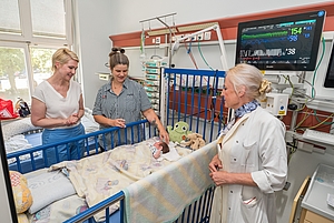 Drei Frauen stehen an einem Krankenhausbett, in dem ein Baby liegt, im Hintergrund medizinische Geräte und ein Fenster