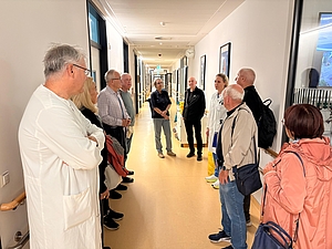 Gruppe von Menschen steht in einem hellen Flur und hört zwei Personen in weißer Arbeitskleidung zu