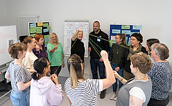 Menschen im Kreis halten Fäden an der Hand, die in der Mitte zusammen führen. 